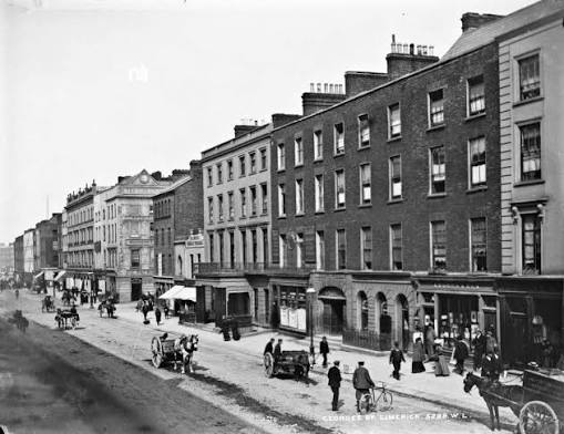 Black-and-white street scene showing O’Connell Street, Limerick, with shopfronts, pedestrians and vehicles.